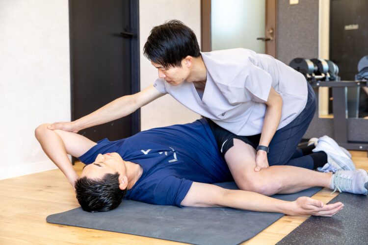 寝転がってストレッチをする男性とサポートするトレーナー