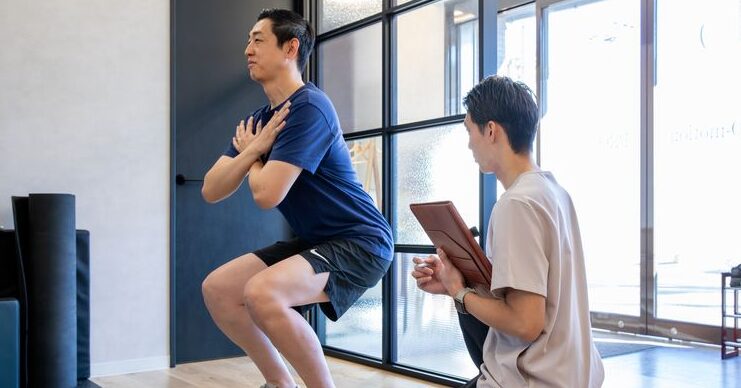 トレーナーが記録を取りながら、スクワット動作を確認している様子